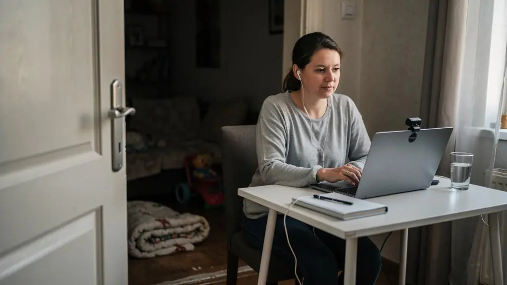 Une femme assise à un bureau dans son salon, portant des écouteurs, participe à une téléconsultation psychologique sur son ordinateur portable, porte fermée.